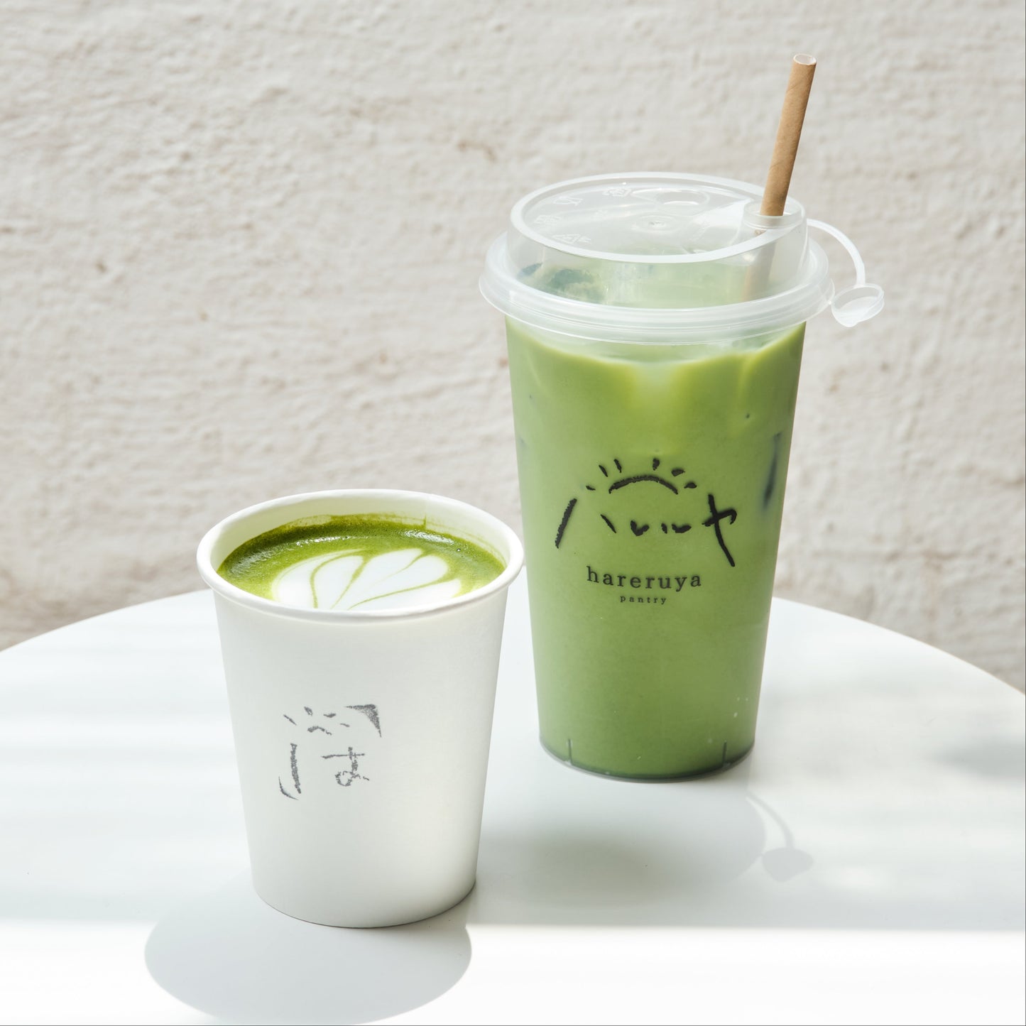 Two cups of matcha green tea, one in a white cup and the other in a clear plastic cup with a straw, on a light surface.