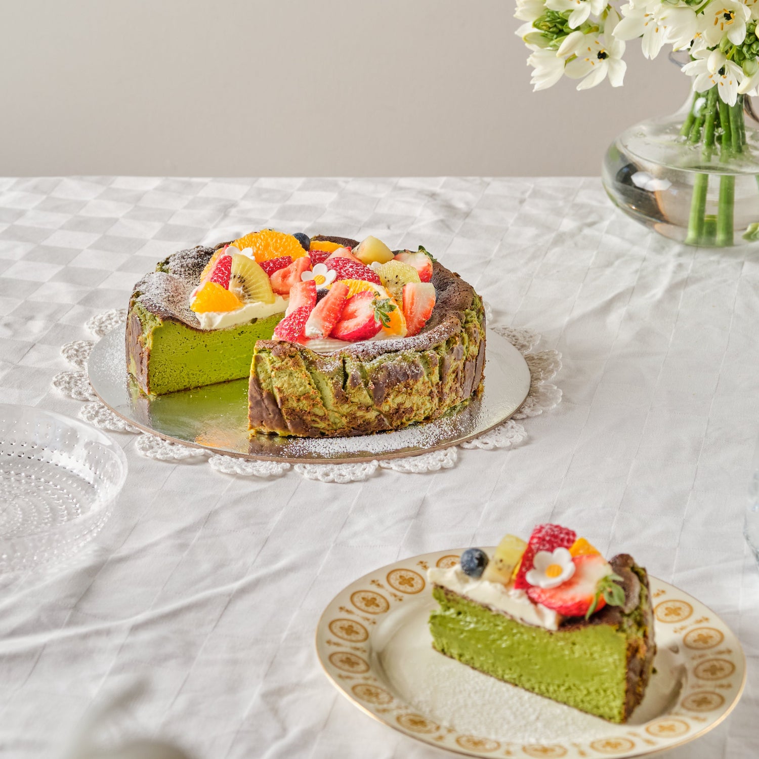 Matcha green tea cake with fruit on a white tablecloth, accompanied by a vase of flowers.